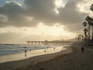 "Panoramic view of Huntington Beach pier on a winter day with moody clouds and golden sunlight, few surfers near the water, people strolling in light jackets, swaying palm trees, and deep blue-green ocean waves under a dramatic partly cloudy sky."