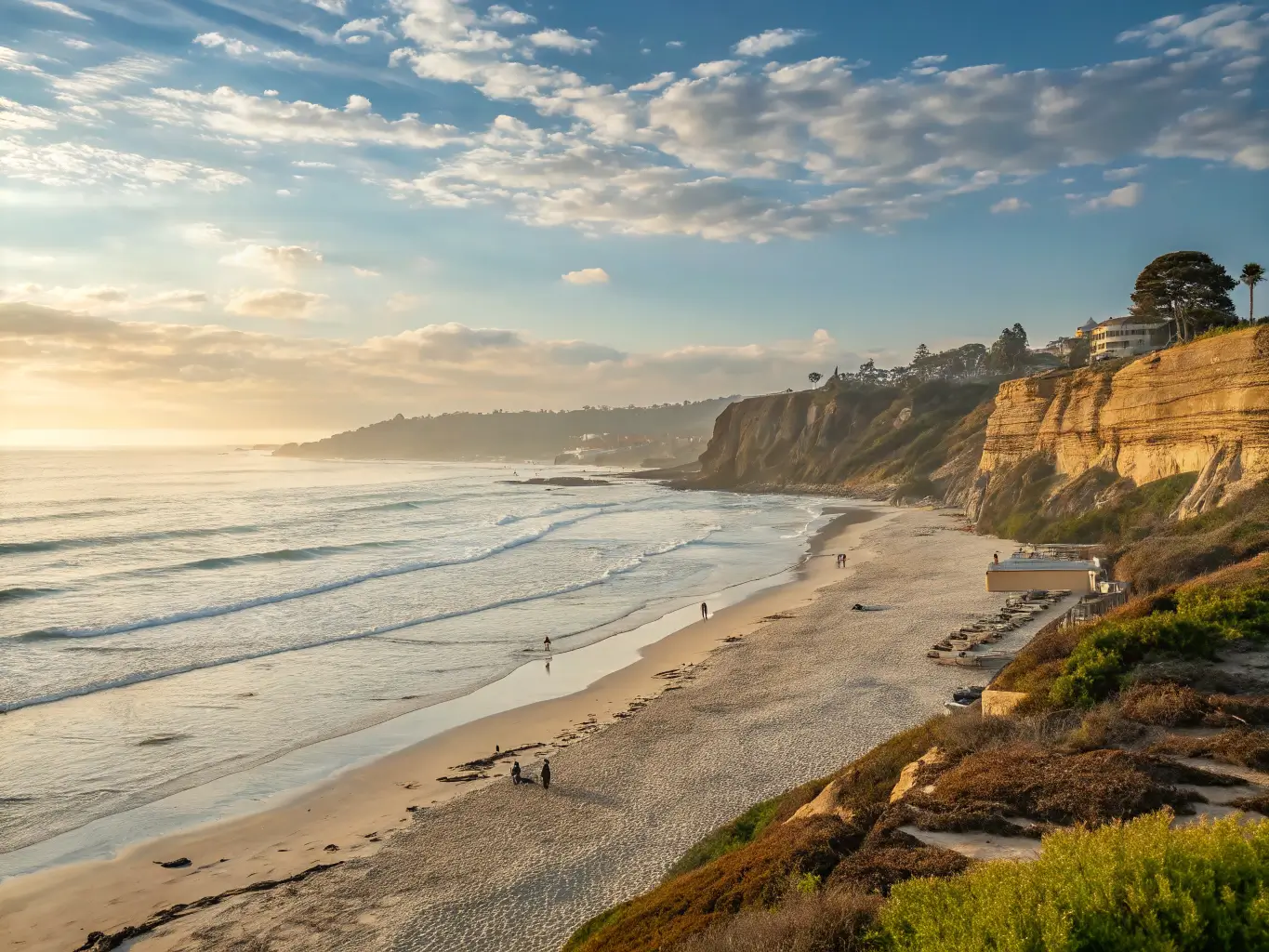 Mid-morning view of La Jolla Shores beach with golden sunlight on calm waves, sandy shore, clear blue sky with wispy clouds, and coastal rock formations in background