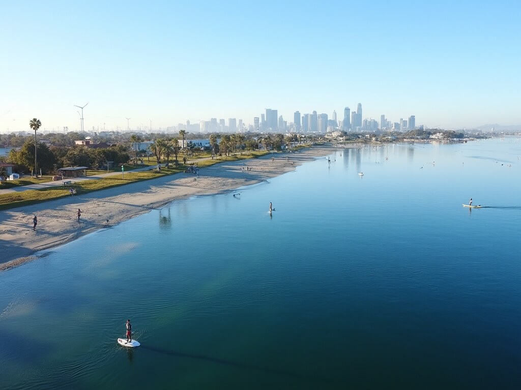 Aerial view of Mission Bay with calm water ideal for paddleboarding, joggers on shoreline paths, and distant San Diego skyline under clear October morning skies