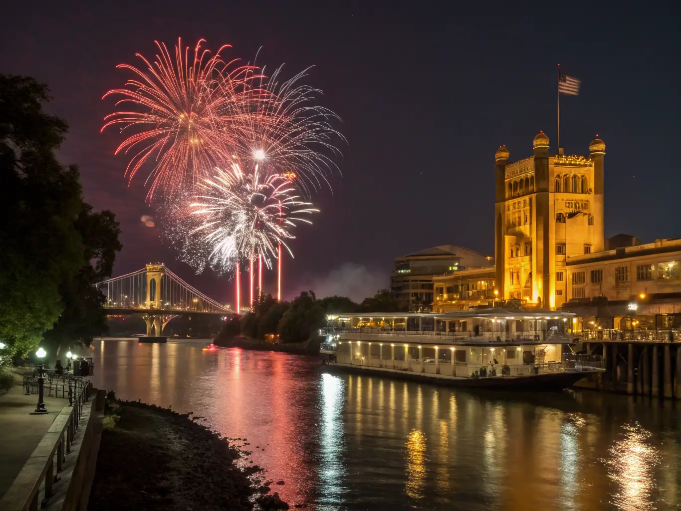 Fireworks display over Old Sacramento Waterfront at night, reflecting in the river, with illuminated historic buildings and the golden glow of Tower Bridge in the background