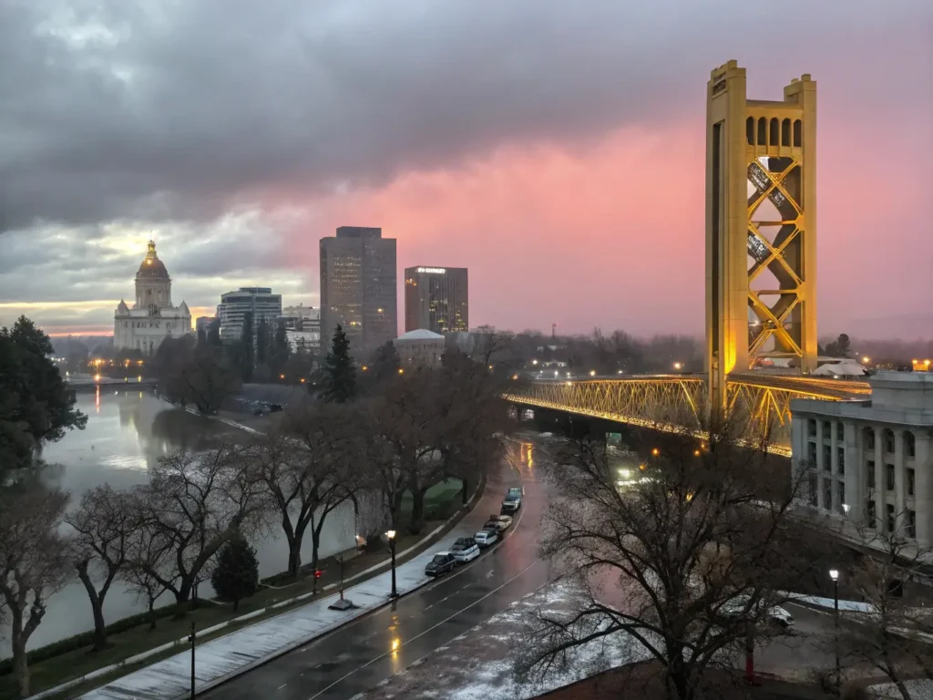 "Downtown Sacramento skyline at dusk with moody rain clouds, pink sunset, golden Tower Bridge reflecting on wet streets, bare winter trees, State Capitol dome, and winter fog."