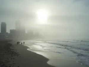 "San Diego skyline obscured by June Gloom fog with sunlight peeping through, empty beach in foreground with a few early morning joggers and surfers, color palette of soft grays and muted blues"