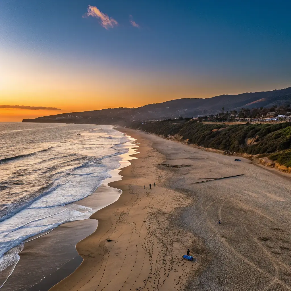 Zuma Beach at golden hour with expansive sandy shoreline, soft waves, clear blue sky and hints of orange sunset