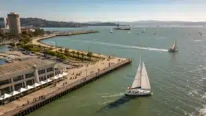 "Aerial view of Jack London Square in Oakland on a sunny August afternoon, showcasing sailboats on the San Francisco Bay, people strolling along the waterfront, outdoor cafes, and the Oakland estuary under a clear blue sky at 72°F."