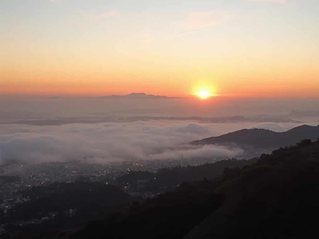 Panoramic sunrise view from Grizzly Peak Boulevard, showcasing the foggy Bay Area with Oakland and San Francisco distant, morning light breaking through layers of fog, and hill and urban silhouette in the backdrop
