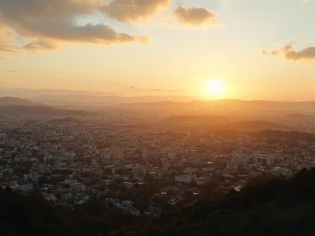 Panoramic view of Oakland cityscape under golden sunlight with urban buildings, distant hills, and clouds filtering soft light, in warm late summer tones, no people visible