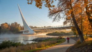 "Autumn view of Redding's Sundial Bridge at golden hour, casting shadows on the Sacramento River, cyclists on riverside trail, surrounded by fall foliage, with morning mist and clear blue sky"