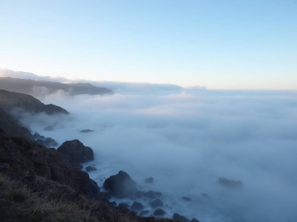 Misty morning on Monterey's Pacific coast, marine layer partially obscuring the coastline and Cannery Row in the distance, transitioning to clear blue sky