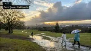 "Scenic view of San Jose, California showcasing layered weather conditions with people in park, winter trees, a visible rain shower, sunny areas, temperature at 54°F, and city skyline in the background"