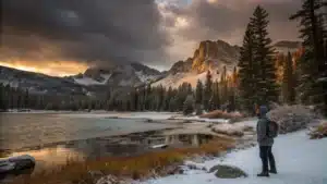 Person in a thermal jacket and beanie stands by a partially frozen alpine lake at Mammoth Lakes at sunset, wind-tousled hair, snow-dusted pines and rocky peaks split between golden light and dark storm clouds; temperature gauge reads 48°F.