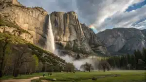 Wide-angle view of Yosemite Falls at peak spring flow plunging down sunlit granite cliffs into spray with a rainbow, green meadow and small visitors in foreground for scale.
