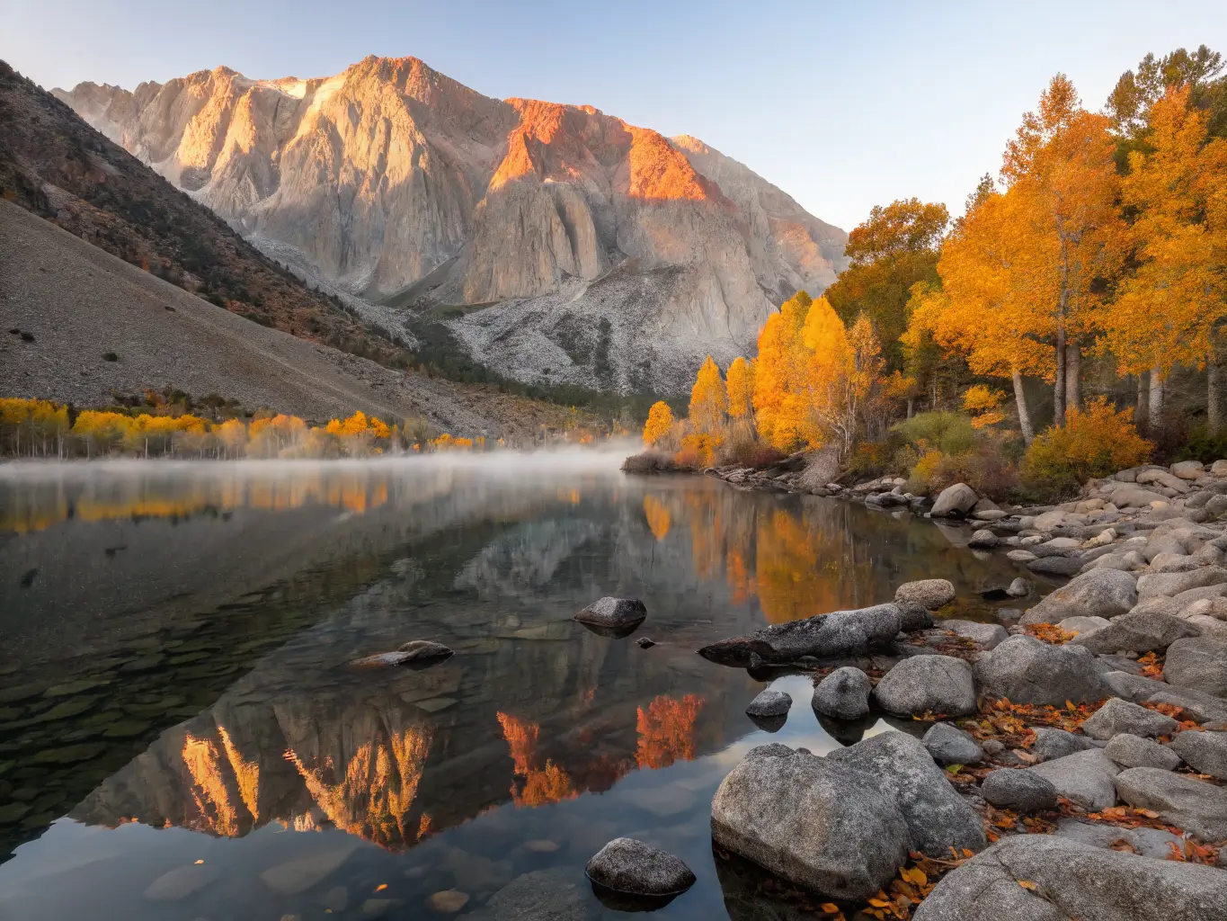 October Might Be the Best Month to Visit Mammoth Lakes Sunrise over Convict Lake in autumn with Mount Morrison reflected in calm blue water, golden aspens along the shore, and foreground boulders with floating yellow leaves.