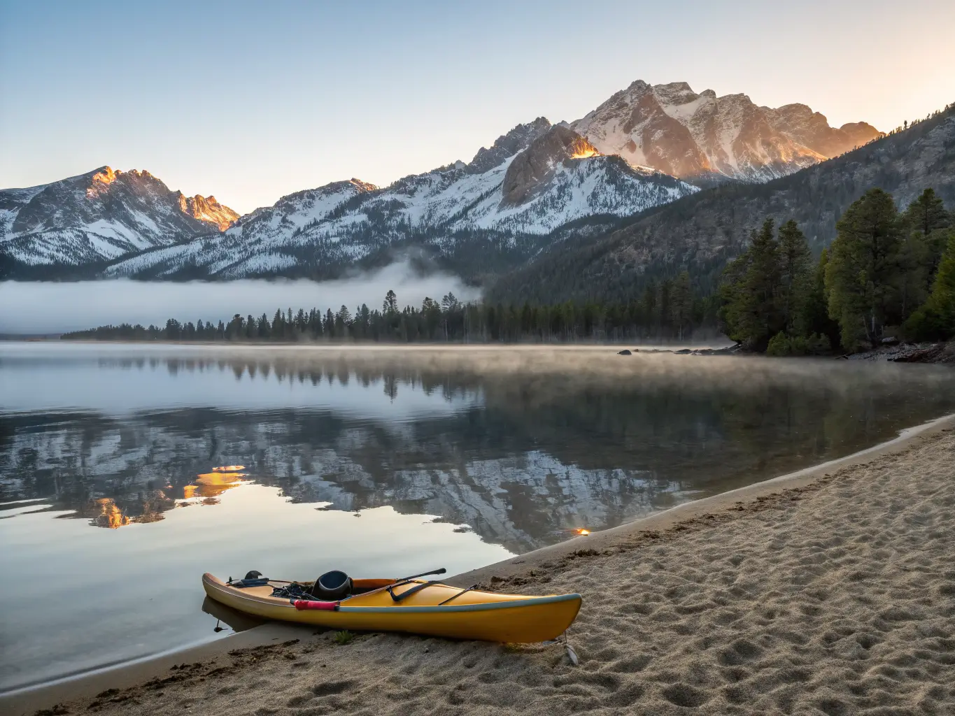 June Might Be the Best Month to Visit Mammoth Lakes Early morning view of June Lake with calm reflective water, snow-capped Sierra peaks, a lone kayak on the sandy shore, and soft golden sunrise light with gentle mist.