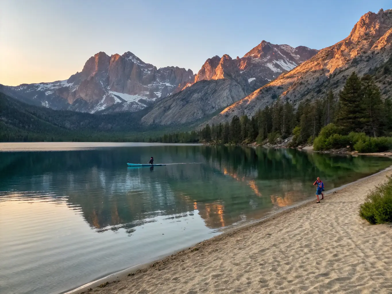 June Might Be the Best Month to Visit Mammoth Lakes Sunrise over June Lake with snow-streaked granite peaks reflected in glassy water, a lone paddleboarder gliding across the calm surface, and sandy turquoise shallows in the foreground.