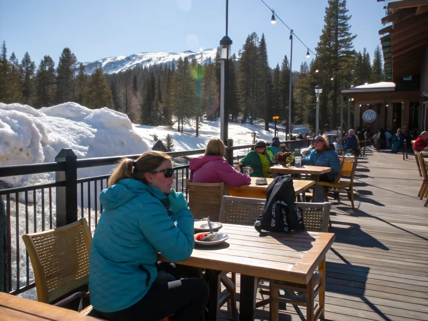Mammoth Lakes in April: What to Actually Expect Skiers relax on a sunny patio in Mammoth Lakes, wearing light jackets and sunglasses, with ski gear nearby and snowy peaks in the background.