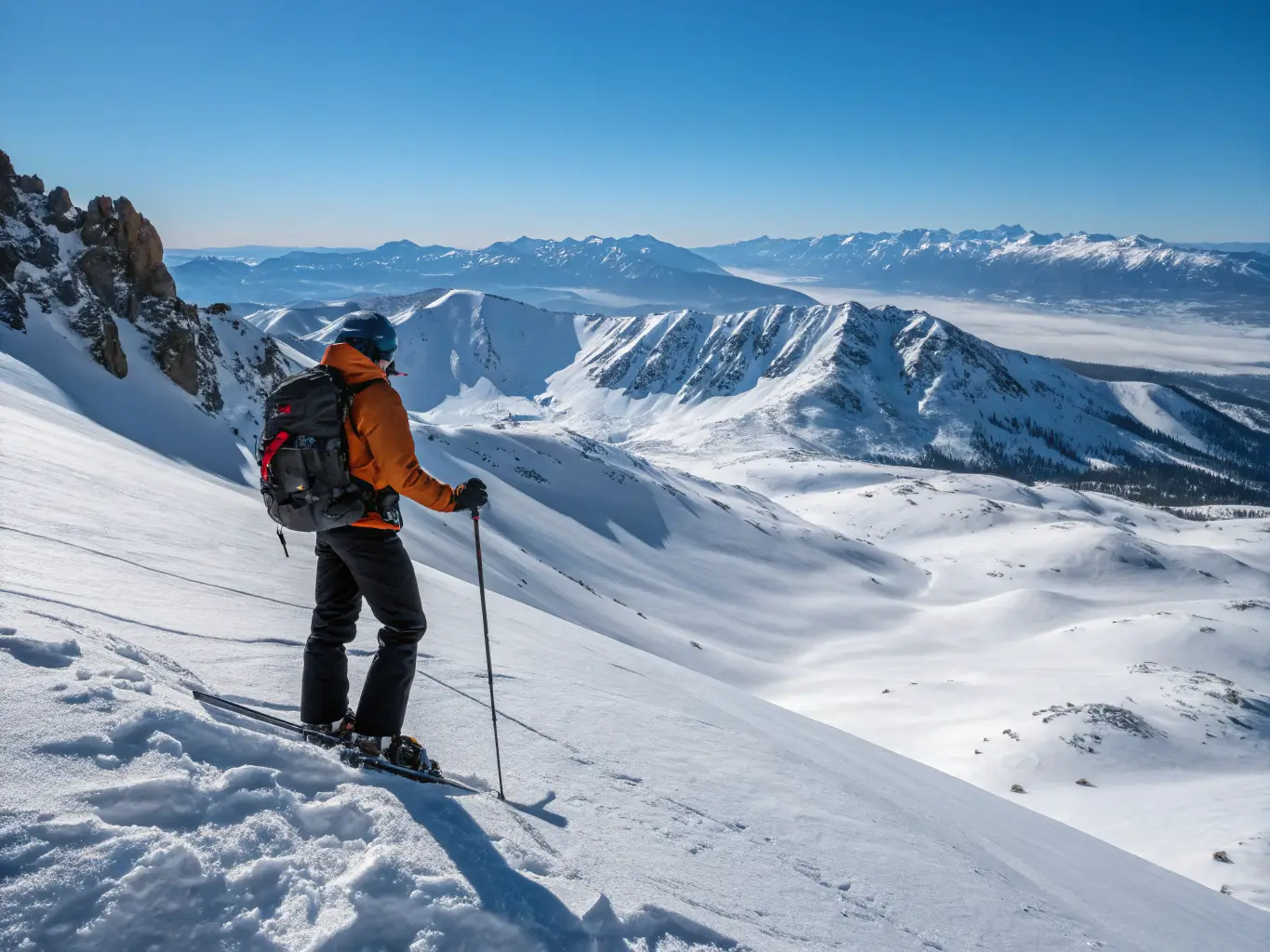 Mammoth Lakes in March: What to Actually Expect Backcountry skier on a high windy ridge near Mammoth summit, avalanche gear visible, overlooking a bright snow-covered Eastern Sierra landscape under a clear blue sky.