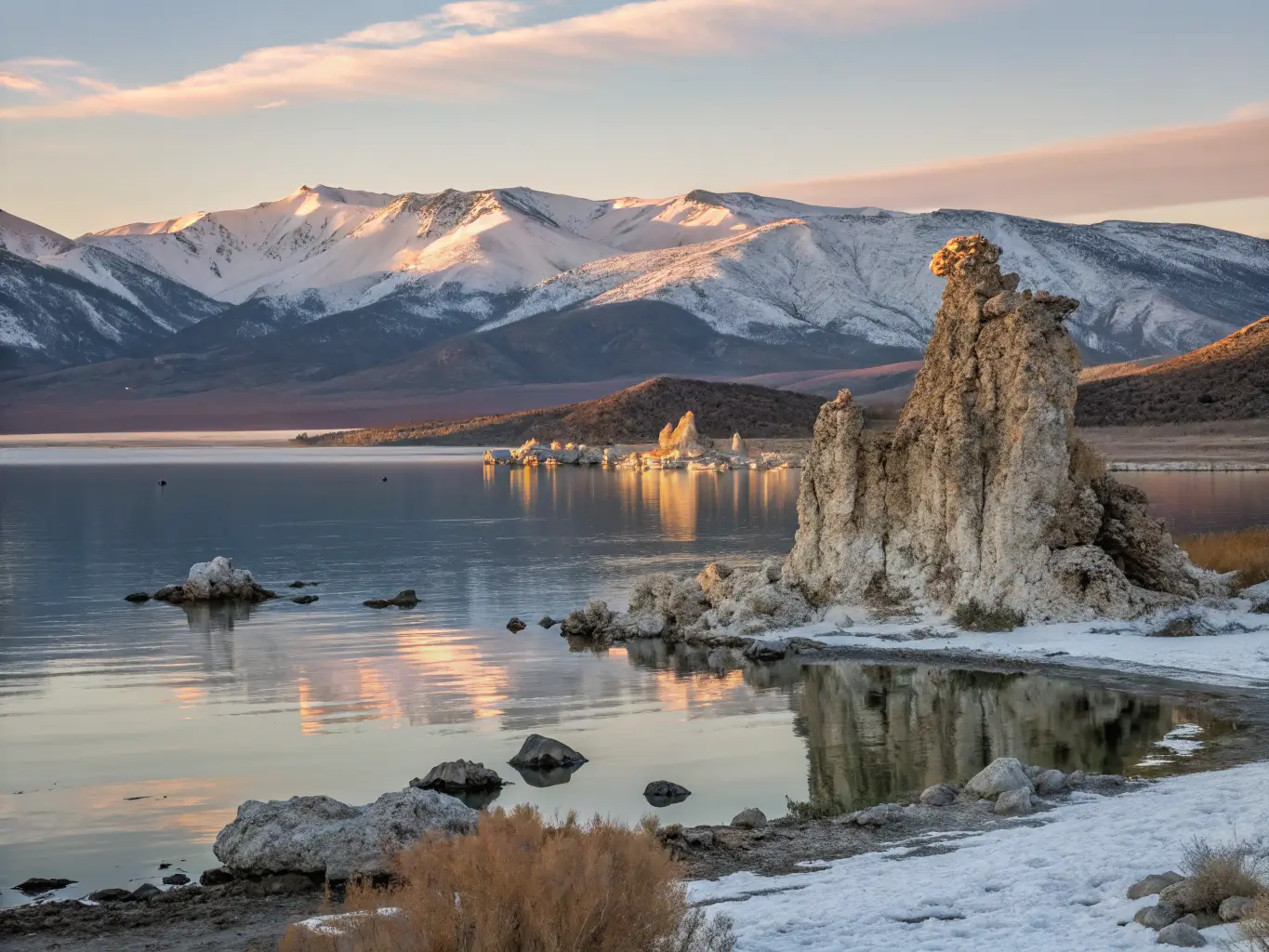 Wide-angle winter view of Mono Lake’s South Tufa at golden hour, showing snow-dusted limestone towers and their perfect reflections on the calm lake, with soft pastel sky and Sierra peaks in the distance.