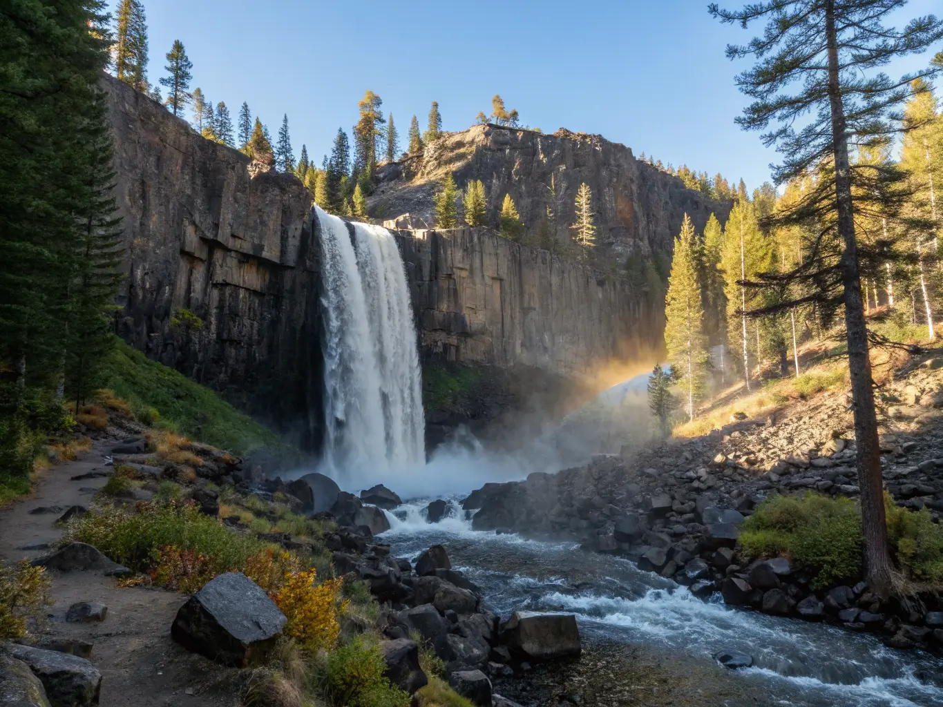 October Might Be the Best Month to Visit Mammoth Lakes Rainbow Falls near Devils Postpile on a clear October morning, waterfall plunging over dark volcanic cliffs with a faint rainbow in the mist, autumn forest and light snow on upper slopes, framed by pine branches.